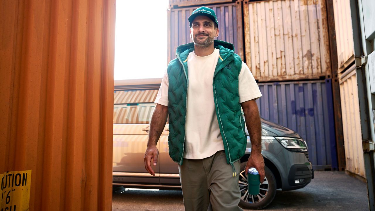 Ein Mann mit Kappe, Weste und Flasche aus der California Collection bewegt sich von einem Volkswagen California fort.