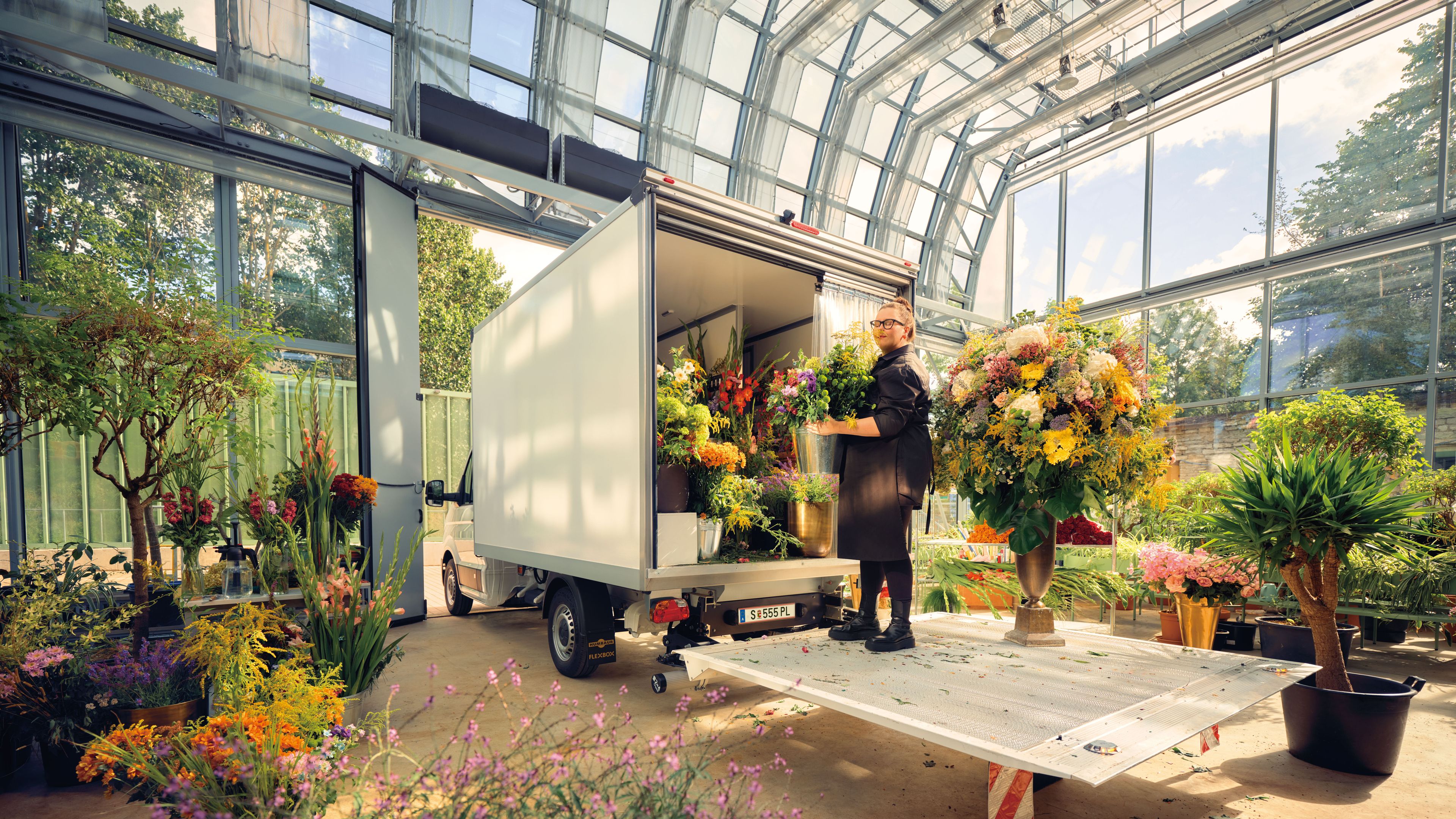 Eine Gärtnerin im Glashaus mit vielen Pflanzen steht auf der Ladefläche eines Volkswagen Transporters.