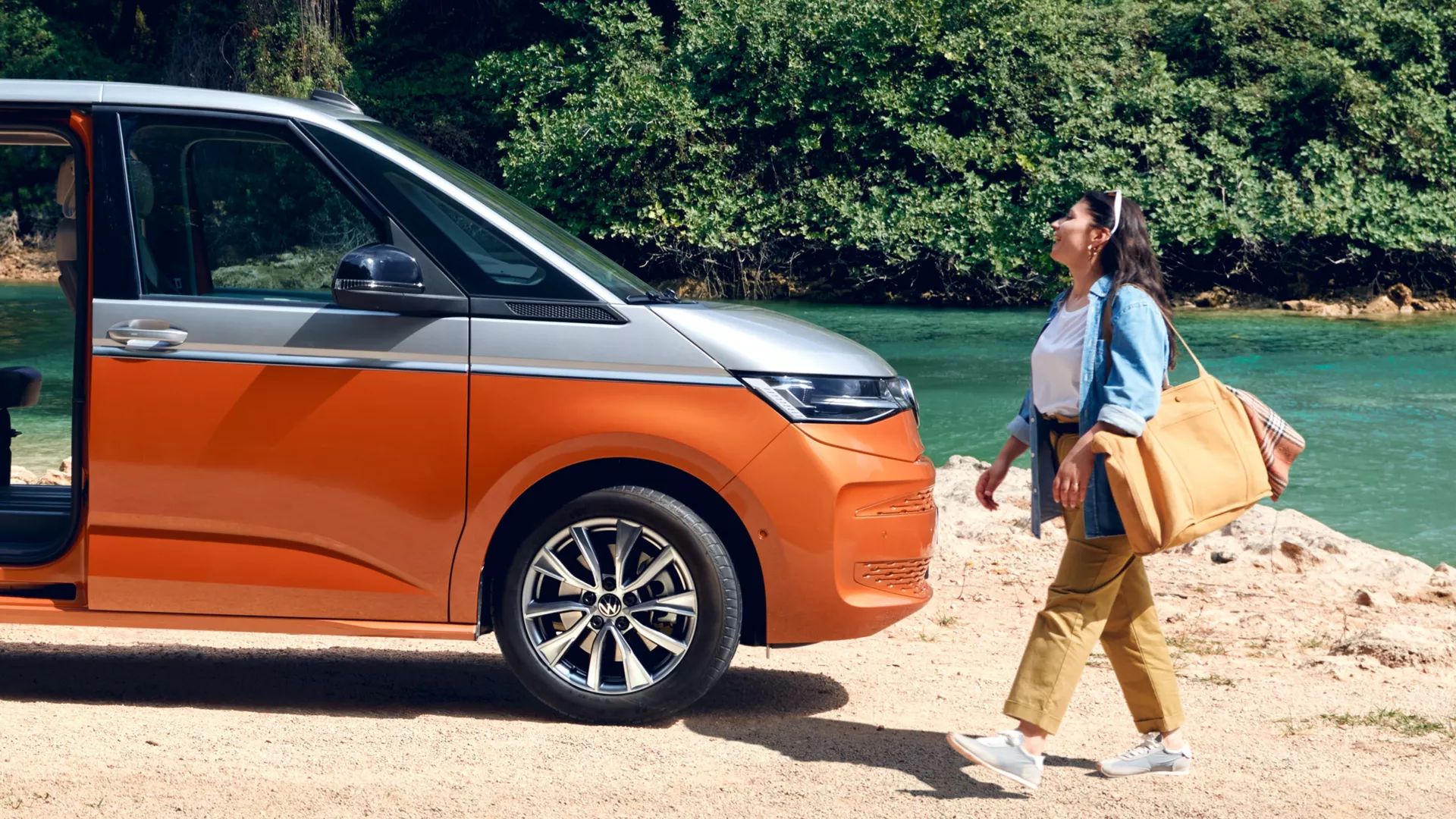 Frau geht auf einen am Fluss parkenden VW Multivan mit geöffneter Schiebetüre zu