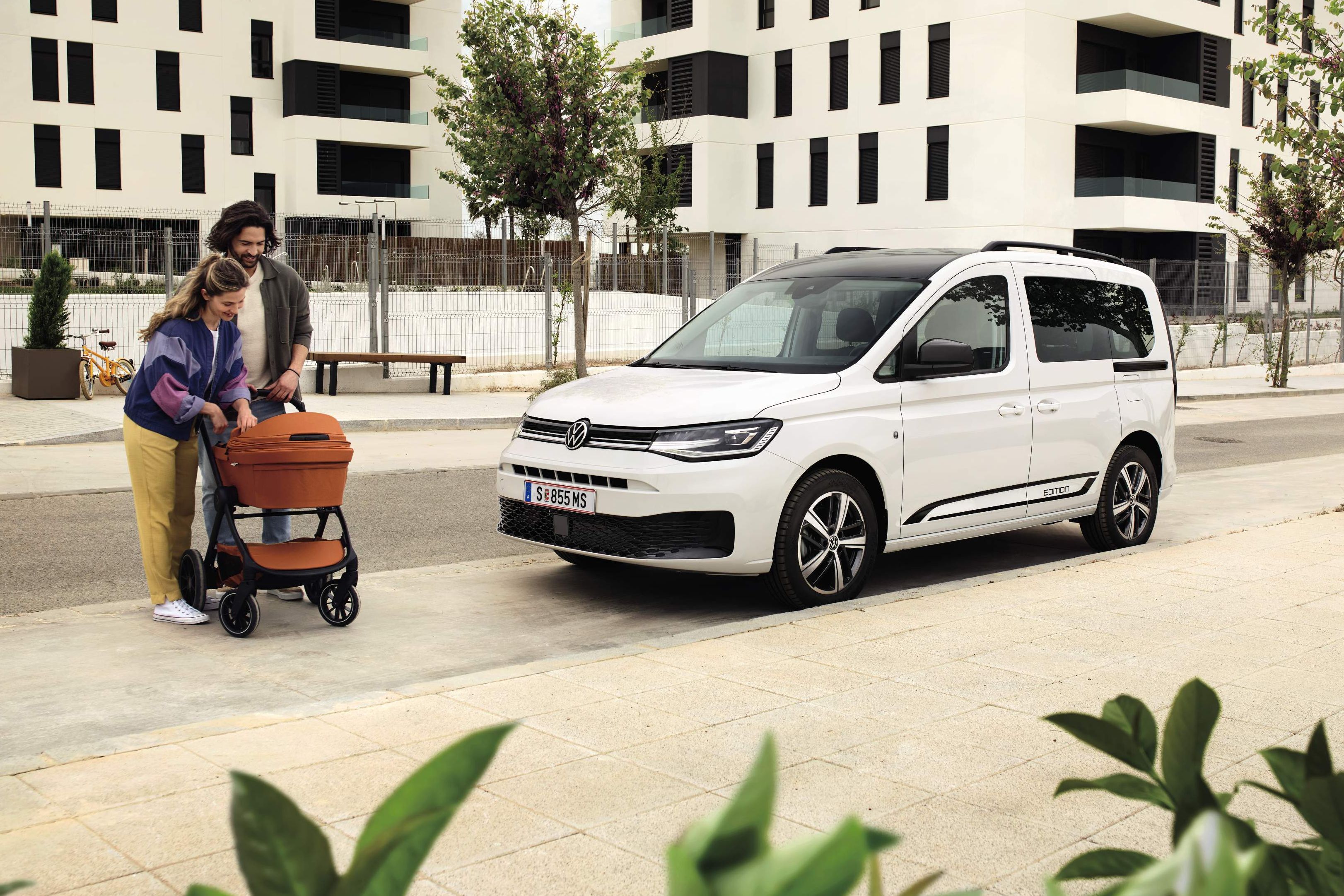 Zwei Erwachsene schieben ein Baby in einem Kinderwagen, Caddy Edition steht daneben