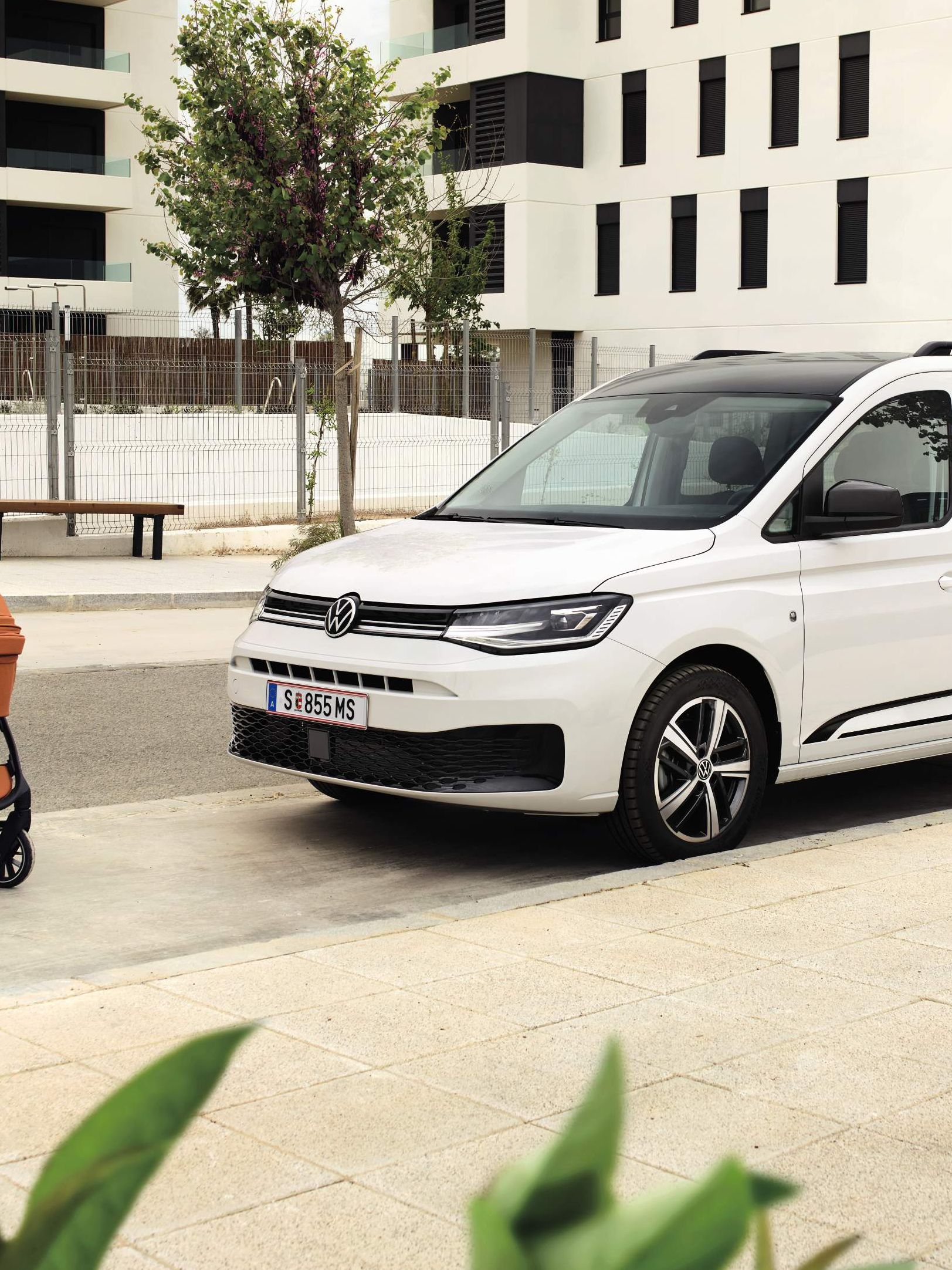 Zwei Erwachsene schieben ein Baby in einem Kinderwagen, Caddy Edition steht daneben