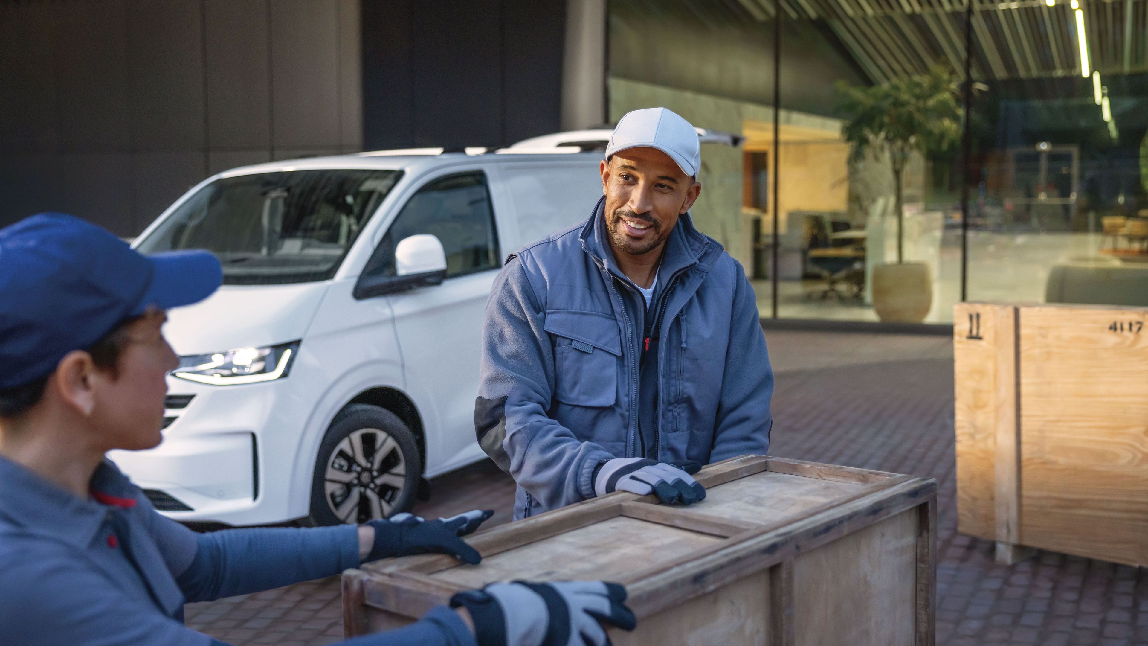 Zwei Männer tragen eine Holzkiste. Im Hintergrund ist ein weißer Volkswagen Transporter zu sehen.