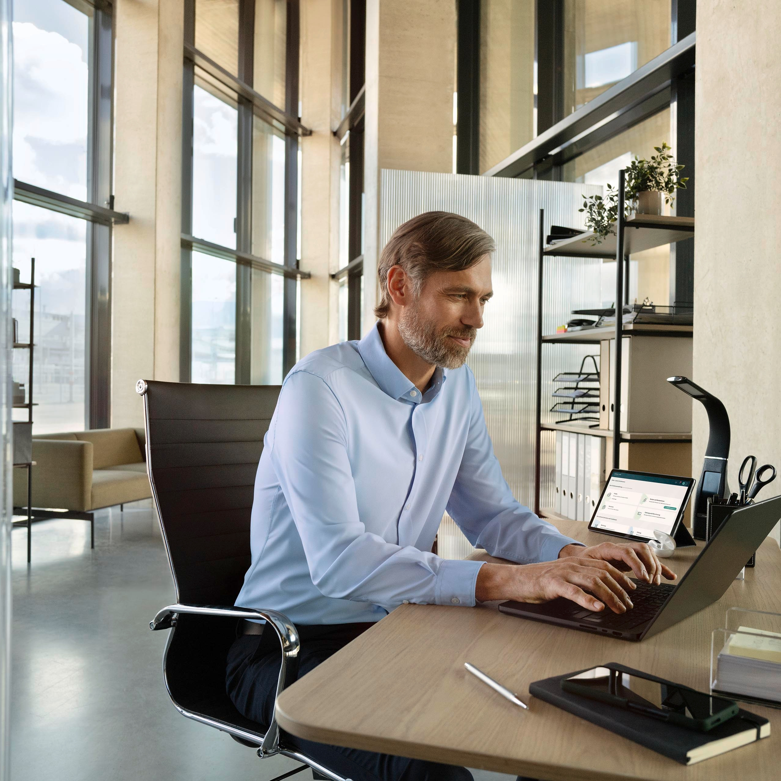 Eine Person sitzt an einem modernen Schreibtisch in einem Büro und arbeitet am Laptop. Im Hintergrund sind große Fenster und Regale mit Büromaterialien zu sehen. Der Raum wirkt hell und gut beleuchtet.
