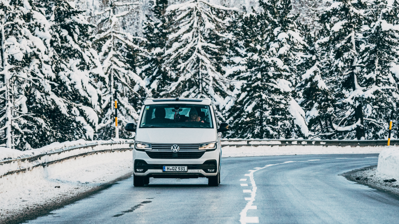 Een weißer Volkswagen Transporter fährt auf einer verschneiten Bergstraße.